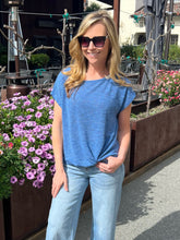 Woman in a blue top and light jeans standing outdoors with flowers and tables in the background