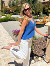 Woman in a blue top and white pants standing outdoors with greenery and a building in the background.