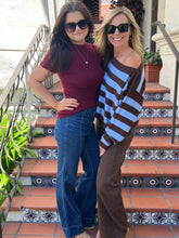Two women posing together on an outdoor tiled staircase. The woman on the left wears a maroon short-sleeve t-shirt, blue jeans, and black sunglasses.