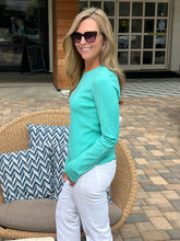 Woman in a teal sweater and white pants standing outdoors near a wicker chair with cushions.