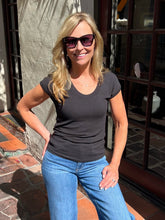 A woman wearing a dark charcoal gray scoop-neck t-shirt and blue jeans, standing outdoors in front of a building with stone walls and glass windows.