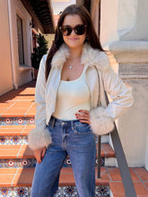 A woman wearing a cream faux-fur trimmed jacket over a cream tank top and blue jeans, standing on outdoor tiled steps.