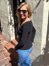 A woman wearing a black knit long-sleeve top with a decorative silver beaded neckline, paired with blue jeans, standing outdoors against a textured stucco wall.