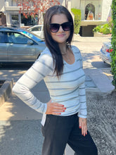 A woman wearing a white long-sleeve top with thin gray horizontal stripes and black pants, standing outdoors on a sidewalk in front of parked cars and a building.