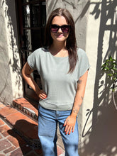 A woman wearing a light sage green short-sleeve knit top and blue jeans stands outdoors against a stucco wall with shadows.