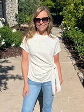 Woman wearing sunglasses and a white t-shirt with a knot at the waist, standing on a sidewalk with greenery.