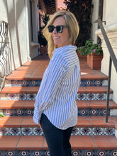 A woman wearing a white long-sleeve button-up shirt with thin blue vertical stripes, paired with black pants and sunglasses, standing on outdoor terracotta steps with tiled accents.