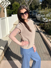 Woman wearing a khaki ribbed knit top standing on a sidewalk with trees and cars in the background