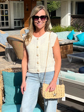 Woman in a cream sleeveless top and light blue jeans standing outdoors with a woven handbag.