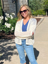 Woman wearing a white crochet cardigan, blue top, and jeans standing outdoors.