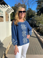 Woman wearing a blue embroidered blouse and white pants standing on a wooden deck.