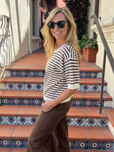 A woman wearing a brown and white striped knit sweater and brown pants stands on tiled outdoor stairs, smiling and wearing dark sunglasses.