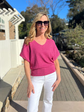 Woman in a magenta sweater and white pants standing outdoors on a sunny day.