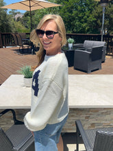 Woman wearing sunglasses and a white sweater with a blue logo outdoors on a patio.