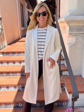 A woman wearing a long cream-colored open-front coat over a black and white striped shirt, styled with dark sunglasses and dark pants, standing on outdoor terracotta tiled steps.
