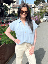 Woman wearing sunglasses and a light blue shirt standing outdoors with greenery and buildings in the background