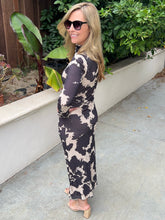 A woman wearing a long-sleeve, high-neck maxi dress with a black and beige abstract cow-print pattern, standing outdoors on a concrete patio next to a planter with green plants.
