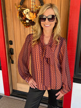 A woman wearing a long-sleeve patterned blouse with a tie-neck detail and black pants, standing in front of a wooden door with a fall wreath.