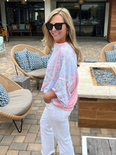 Woman in a pink patchwork top and white pants standing outdoors near patio furniture.
