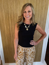 Woman wearing a black sleeveless top and patterned pants against a gold leaf wall.