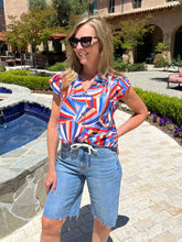 Woman wearing a colorful shirt and denim shorts standing outdoors near a pool.