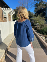 Woman wearing a blue embroidered blouse and white pants standing on a wooden deck.