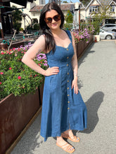 Woman in a dark chambray midi dress standing on a sidewalk with flowers and buildings in the background