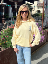 Woman wearing a light yellow hoodie with an anchor design, standing outdoors near flowers and plants.