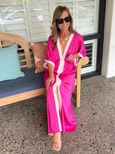 Woman in a pink dress with white trim sitting on a wooden bench outdoors.