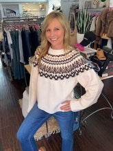 A woman wearing a cream knit sweater with a brown and beige geometric Fair Isle yoke pattern, paired with blue jeans, posing inside a clothing boutique.