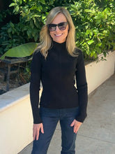 A woman wearing a black ribbed mock neck long-sleeve top and dark blue jeans, standing outdoors in front of a beige wall and green foliage.