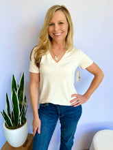 Woman wearing an ivory V-neck t-shirt and blue jeans standing next to a potted plant against a light blue wall.