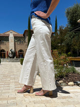 Person wearing white pants and a blue top standing on a stone path with a building and trees in the background.