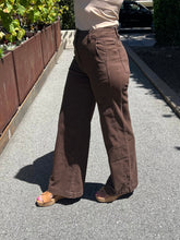 A person wearing brown wide-leg corduroy pants and tan slide sandals, standing on an outdoor paved pathway.