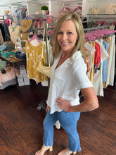 Woman wearing white blouse and h jeans standing in a clothing store with racks behind her.