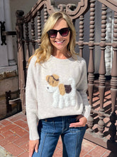 A woman wearing a cream-colored knit sweater with a raised dog motif on the front, paired with blue jeans and dark sunglasses, standing outdoors in front of a decorative wooden railing.