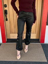A person wearing black flared pants with decorative metal button details down the outer calves, paired with a burgundy sweater and beige heels, standing on a doormat in front of a wooden door.