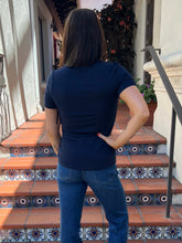 A woman viewed from behind wearing a navy blue short-sleeve t-shirt and blue jeans, standing on terracotta tiled steps with decorative blue and white patterned tiles.