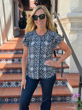 A woman wearing a short-sleeve button-front blouse with a black and white geometric floral pattern, paired with dark blue jeans and sunglasses, standing on outdoor terracotta tile steps.