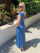Woman in a blue patterned dress standing outdoors near plants.