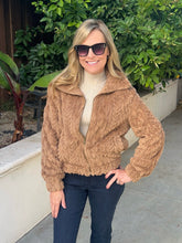 A woman wearing a brown textured zip-up jacket over a cream turtleneck and dark blue jeans, standing outdoors in front of green plants and a concrete wall.