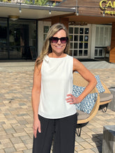 Woman in a white sleeveless top and black pants standing outdoors with a building in the background.