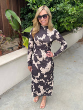 A woman wearing a long-sleeve mock-neck midi dress with a black and beige abstract cow print pattern, standing outdoors on a concrete walkway with green plants and a wooden fence in the background.