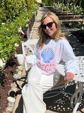 Woman sitting on a white bench in a garden wearing a white sweatshirt with a blue and pink design.