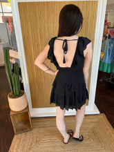 Person wearing a black dress with ruffled sleeves and open back, standing in front of a wall.