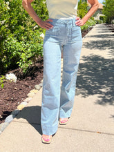 Person wearing light blue jeans standing on a sidewalk with greenery in the background