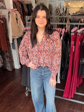 A woman with dark hair stands in a clothing boutique wearing a long-sleeve V-neck blouse with a multicolor abstract pattern in orange, blue, and pink, paired with light-wash blue jeans.