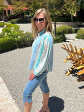 Woman wearing sunglasses and a light-colored jacket standing outdoors with greenery and a wooden chair in the background.