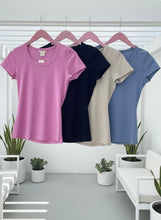 Four t-shirts in pink, black, mocha, and denim hanging on a rack with plants in the background.
