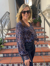 A woman wearing a long-sleeve V-neck blouse with a blue, purple, and orange abstract pattern, paired with dark jeans and sunglasses, standing on terracotta tiled stairs with decorative blue and white mosaic tiles.